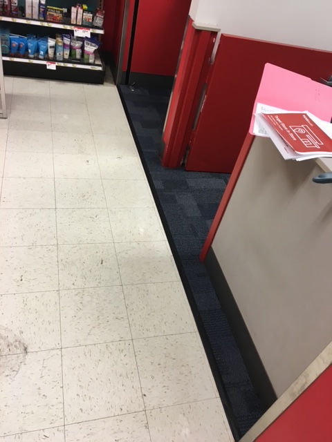 A retail store floor transition showing white tiles on the left meeting dark carpet by a red checkout counter.