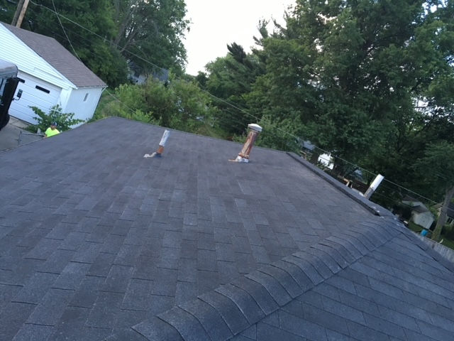 A dark-shingled roof viewed from above, featuring two protruding plumbing vent pipes and a house and trees in the background.