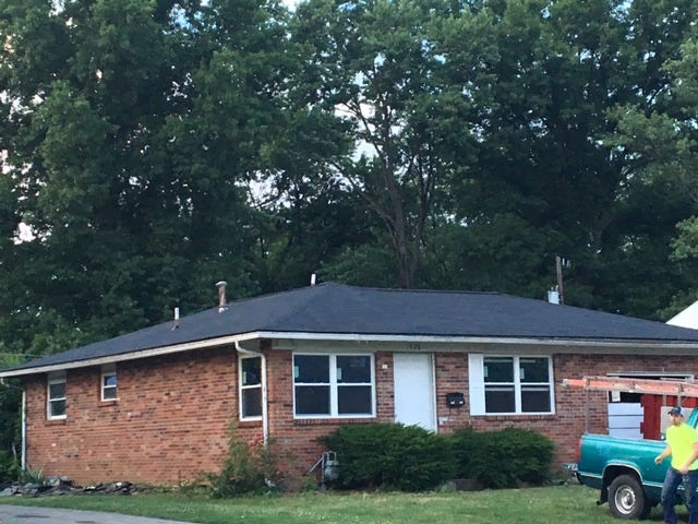 A single-story brick house with a new dark shingle roof stands near a turquoise truck parked on a grass lawn.