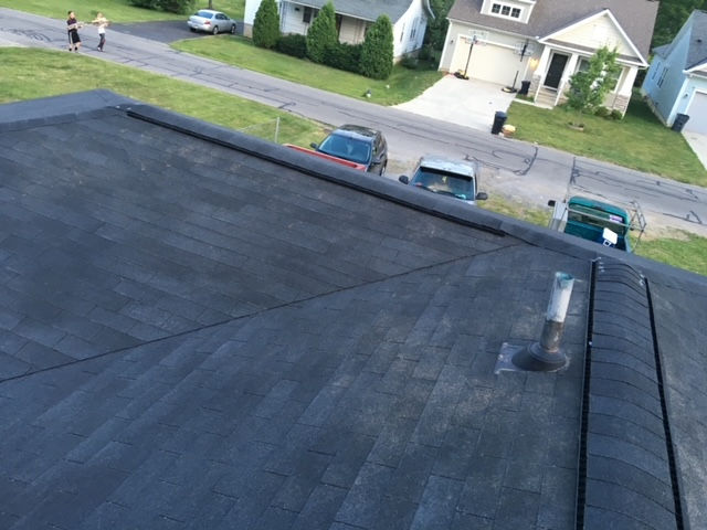 A high-angle view of a dark asphalt shingle roof with a vent pipe, overlooking a residential street and houses.