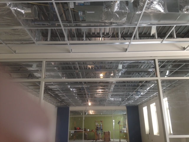 Interior view of a modern office space under renovation, showing exposed ceiling grid, glass walls, and a green accent wall.