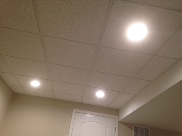 A view of a white drop ceiling with three recessed LED lights, situated above a beige wall and a door frame.