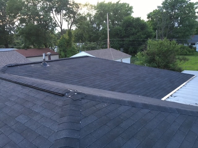 A view from above looking across dark asphalt shingles on a low-sloped residential roof towards surrounding trees.