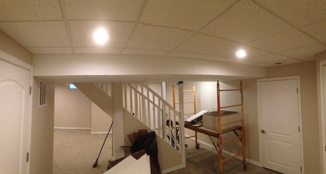 An interior view of a basement renovation with a central support beam, a staircase, and scaffolding near a white door.