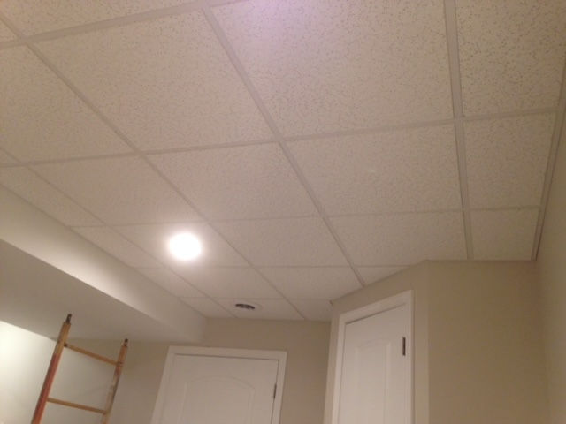 A white drop ceiling with a recessed light fixture and a partially visible ladder in a room with beige walls and doors.