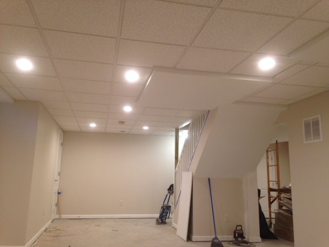 A basement interior featuring beige walls, a drop ceiling with recessed lighting, and a staircase leading to another level.