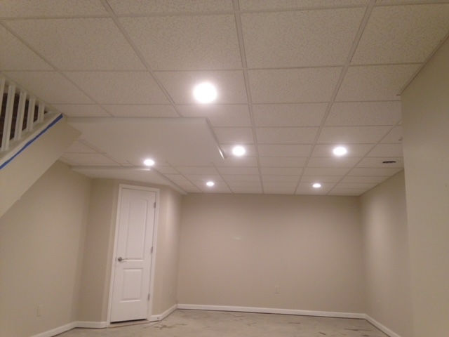 A finished basement room with a white drop ceiling featuring recessed lights, beige walls, and a white closed door.