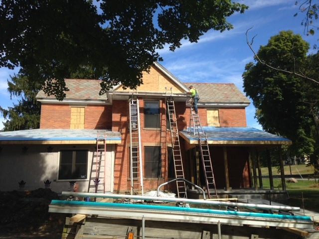 Workers on ladders replace gutters on the roof of a two-story red brick house under renovation.