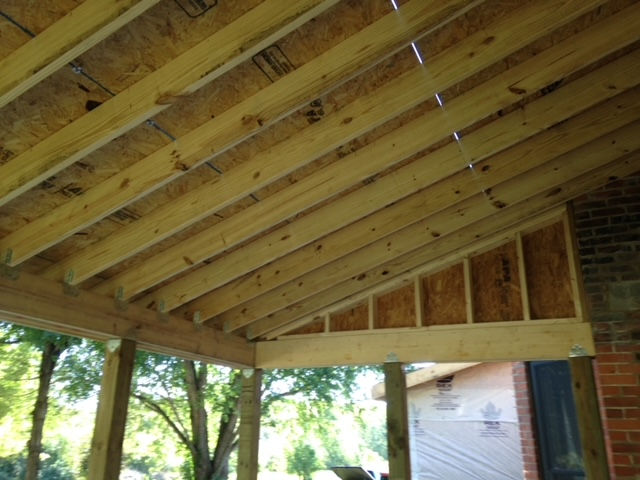 Underside view of wooden porch rafters and joists under construction, attached to a brick wall.