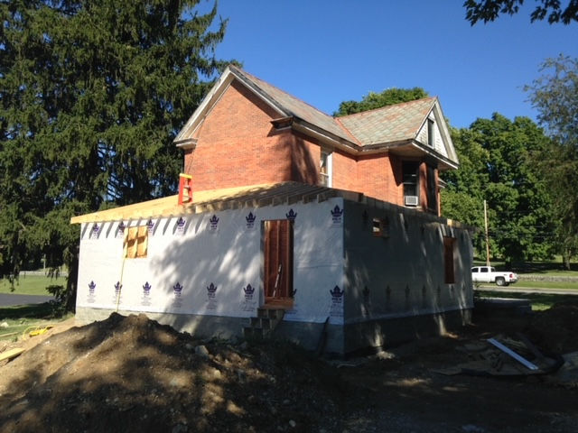 A historic brick house under renovation with a new, unfinished ground-floor addition wrapped in white house wrap.