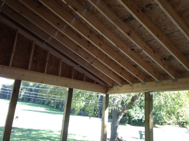A view looking up at the exposed wood framing, rafters, and roof decking of an unfinished outdoor porch structure.