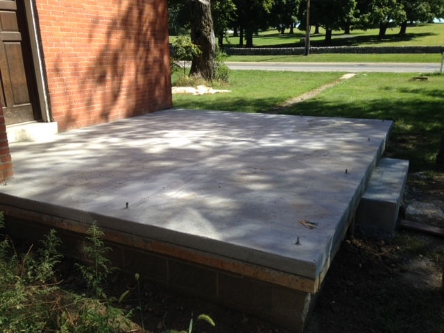 A new concrete slab porch is built against a brick building, elevated above ground level with block supports.