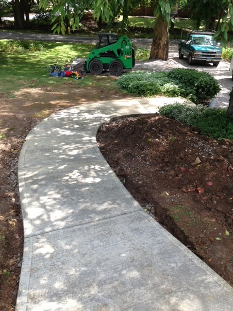 A curved concrete walkway leads through a landscaped yard, with a green skid steer and a truck parked in the background.