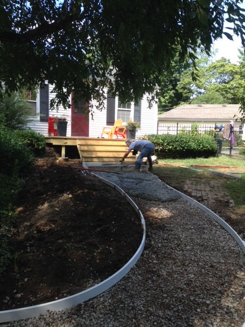 A person works on a gravel path leading to new wooden deck stairs at a house with white siding and a red door.