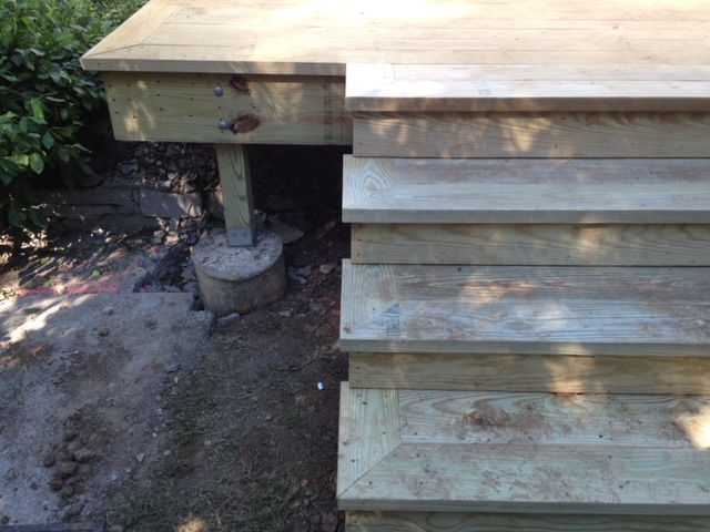 Wooden deck steps leading down from a deck platform to dirt ground, supported by a concrete pier.