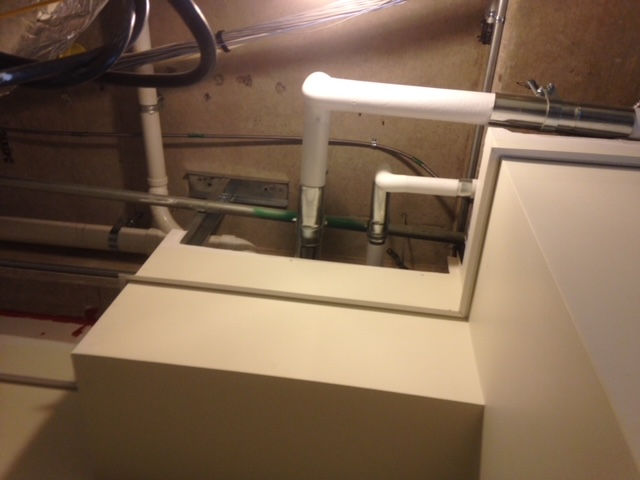 White PVC plumbing pipes extend from an opening in a beige, box-like enclosure near a wall.