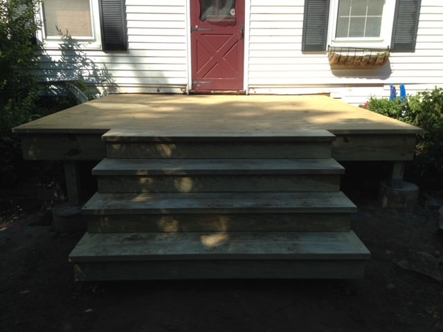 A wooden deck with four stairs leading up to a house with a red door and white siding.