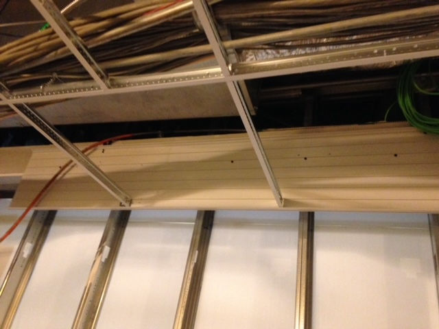 A construction site showing metal wall studs, a wooden board installation, and a ceiling grid with exposed wiring.