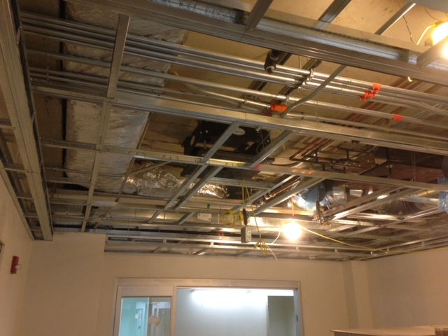 Exposed metal ceiling grid framework under construction, revealing wires, pipes, and ventilation ducts above.