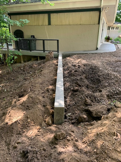 A concrete footing for a retaining wall sits in a dirt yard next to a house with a deck.