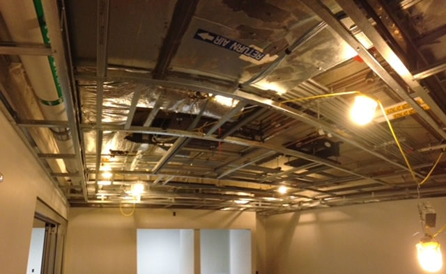 A view looking up at a metal ceiling grid under construction, with exposed pipes, wiring, and temporary hanging lights.