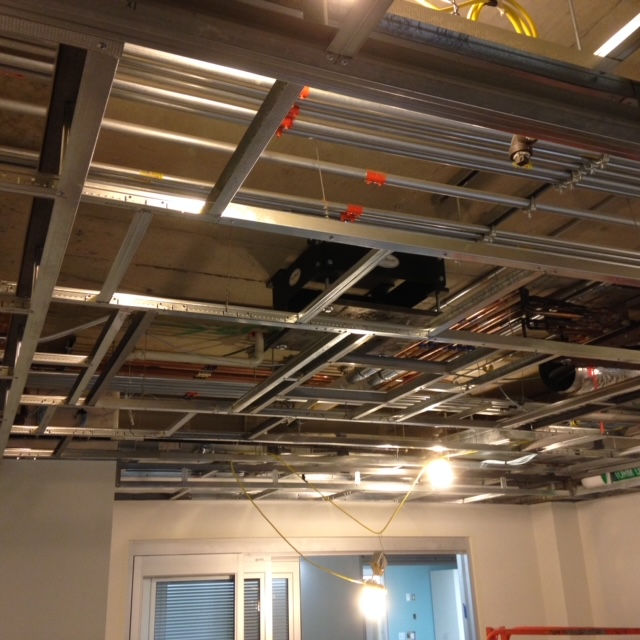 A metal frame for a suspended ceiling under construction, with exposed pipes and wiring against a concrete ceiling.