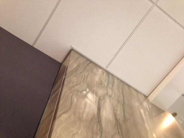 A low-angle view showing the corner junction of a grey wall, a wood-patterned surface, and a white drop ceiling.