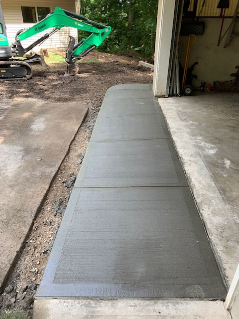 A freshly poured concrete ramp leading into a garage, with a green excavator parked on the dirt lot in the background.