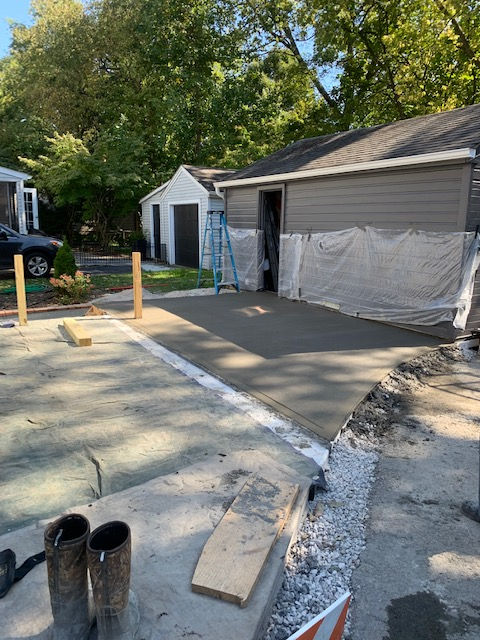 A freshly poured concrete driveway section next to a garage with a white plastic tarp covering the siding.