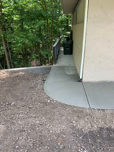 A new concrete walkway curves alongside a beige building wall next to a wooded area and a green trash bin.