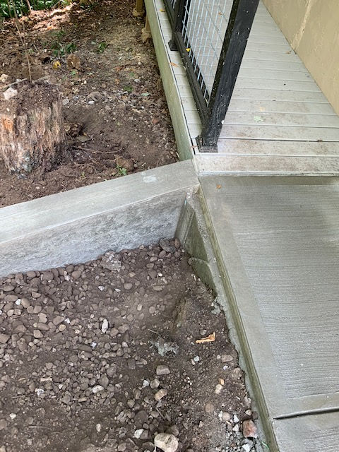 A gray concrete ledge creates a transition between a dirt yard and a concrete walkway near a railing.