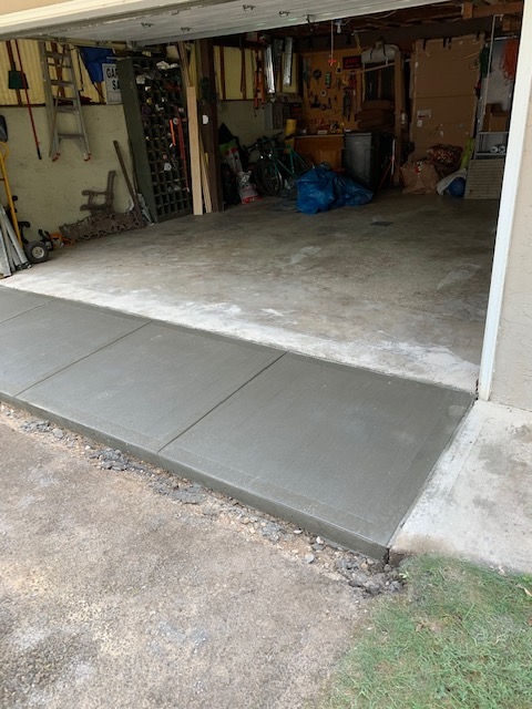 A newly poured, smooth gray concrete transition strip between an old asphalt driveway and a dark, cluttered garage interior.