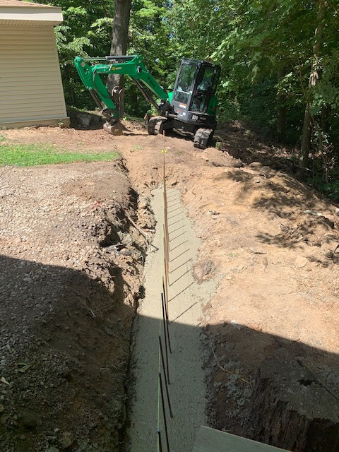 A green mini-excavator parked near a freshly poured concrete footing trench with vertical rebar set in the ground.