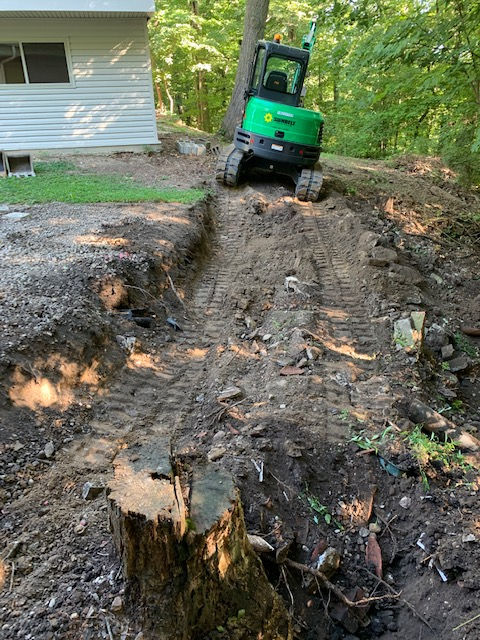 A green mini excavator moves through a dirt path beside a white house near a wooded area.