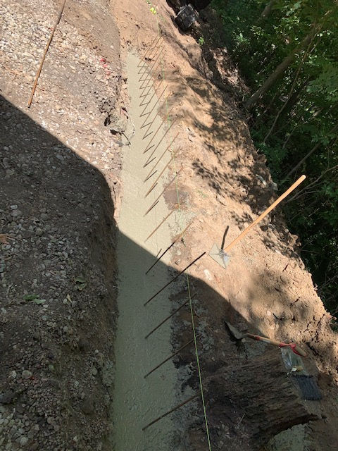 Fresh concrete foundation footing in a dirt trench with vertical rebar dowels protruding along the center.