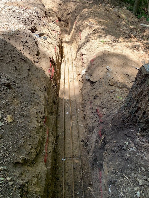 A narrow trench dug into dirt, containing multiple long, parallel metal rebar rods laid along the bottom for construction.