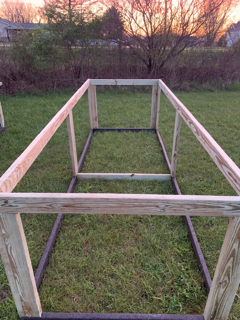 A wooden rectangular frame stands on a grassy lawn during sunset, likely a base for a garden bed or animal enclosure.