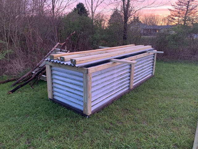 A rectangular raised garden bed made of corrugated metal and wooden framing, sitting on a grassy lawn at sunset.