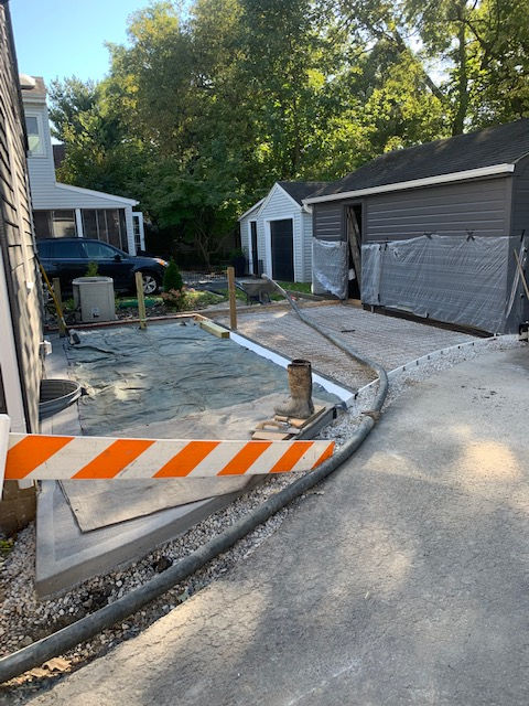 A backyard construction site shows a newly poured concrete patio foundation, a gravel base, and a grey garage with a tarp.
