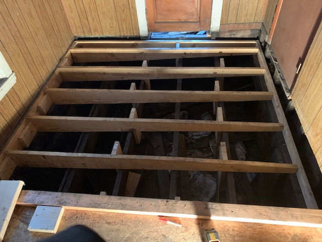 A renovation project showing exposed wooden floor joists and support beams inside a room with wood-paneled walls.