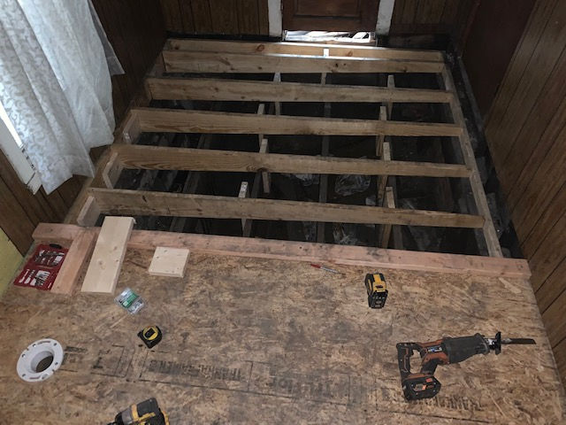 A view looking down at a wooden floor joist structure under construction, with tools and a toilet flange on subflooring.