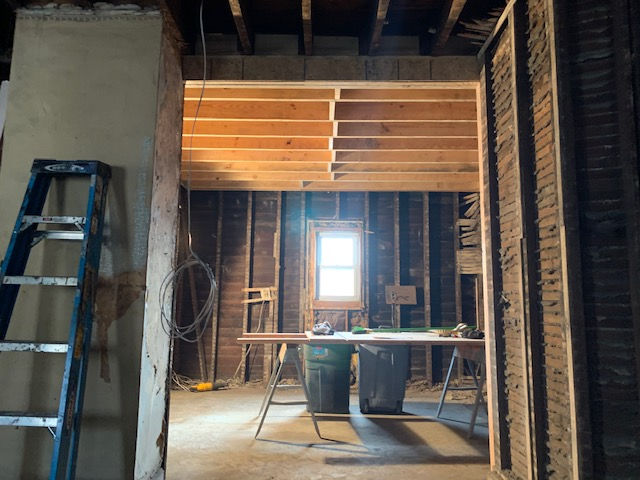 An unfinished room under renovation with exposed wooden joists, wall studs, lath, a blue ladder, and a central window.