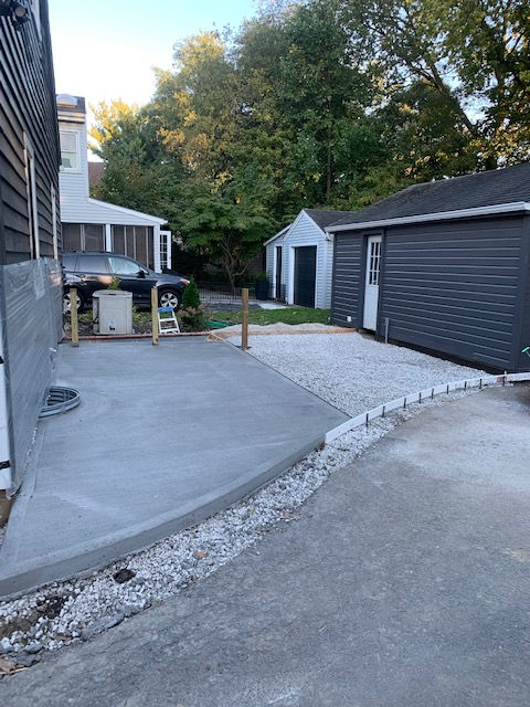 A new concrete patio with a gravel border next to a dark gray garage and a house, with a car parked in the background.