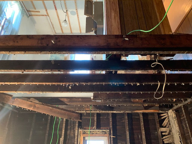 Interior view of a room under renovation, showing exposed wooden floor joists, wall framing, and hanging electrical wires.