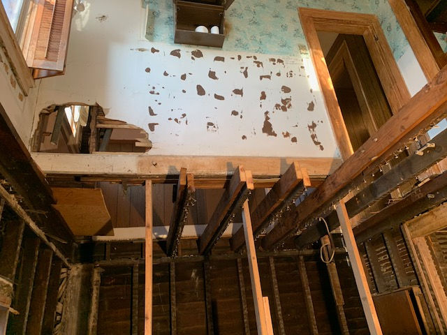 A renovation project in progress showing exposed floor joists, wooden supports, and a wall with damaged plaster.