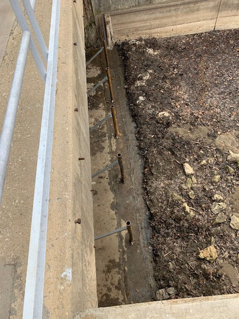 A concrete wall with metal support rods extending into an adjacent dirt and gravel embankment.