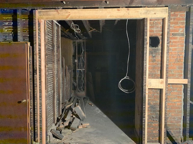 A construction frame for a doorway stands in a dark, unfinished room with exposed brick walls and debris on the floor.
