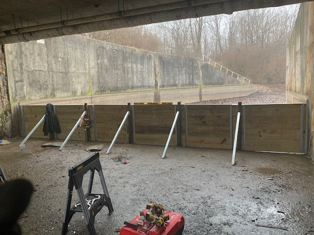 A wooden flood barrier braced by angled metal supports stands inside a concrete structure, with a forest in the background.