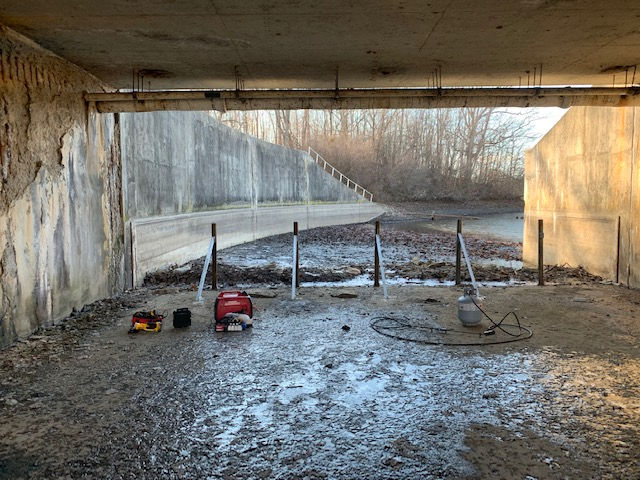 A red generator, tool bags, and a pressurized water hose sit under a concrete overpass near a flooded, muddy bank.