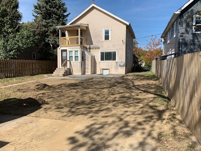 A two-story beige house with a wooden back deck, overlooking a dirt yard bordered by wooden fences on a sunny day.